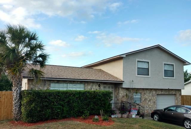 Tan home with brick & siding, brown roof, yard has a palm tree, grass & hedges