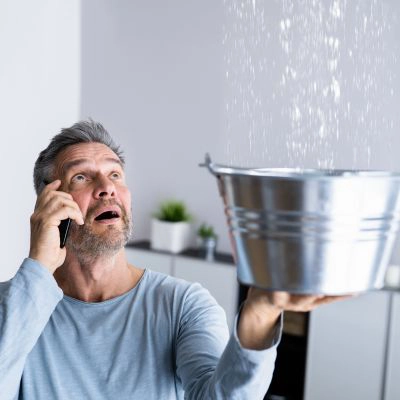A man stands in his house holding a bucket, catching water from a roof leak