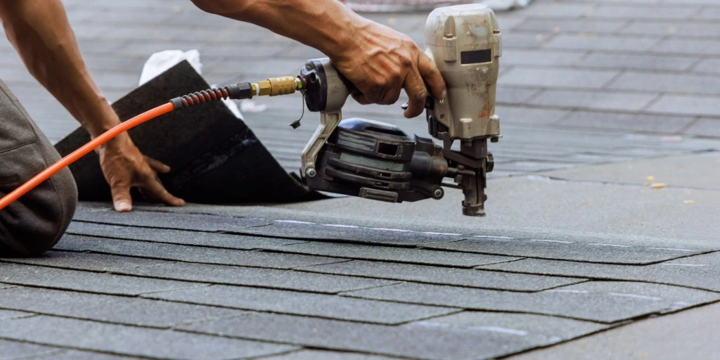Professional roofer using pneumatic nail gun to install gray asphalt shingles on residential roof