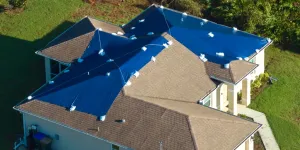 Aerial view of residential Florida home with blue emergency tarps covering damaged roof sections after hurricane, showing multiple roof slopes requiring storm damage repair assessment for 25% rule calculation