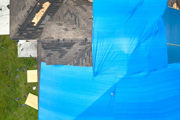 Aerial view of a storm-damaged roof with a blue emergency tarp covering missing shingles and exposed decking after a severe weather event